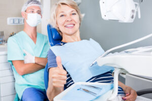 Smiling woman giving a thumbs up in dental chair after successful treatment with dentist in background.